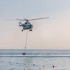 A helicopter with a red basket descends over the sea to scoop water against the background of the dawn orange sky and the silhouette of the city in the distance