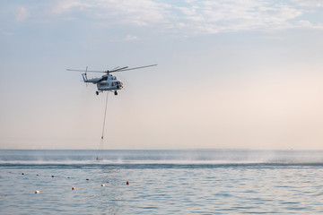 A helicopter with a red basket descends over the sea to scoop water against the background of the dawn orange sky and the silhouette of the city in the distance
