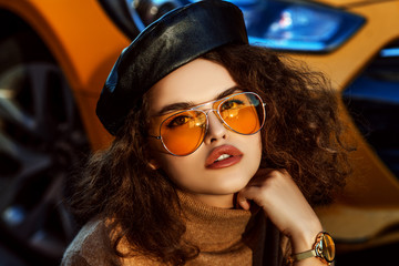 Outdoor close up fashion portrait of young beautiful confident woman wearing stylish orange aviator sunglasses, leather beret, cashmere turtleneck,  wrist watch, posing in street, near the bright car