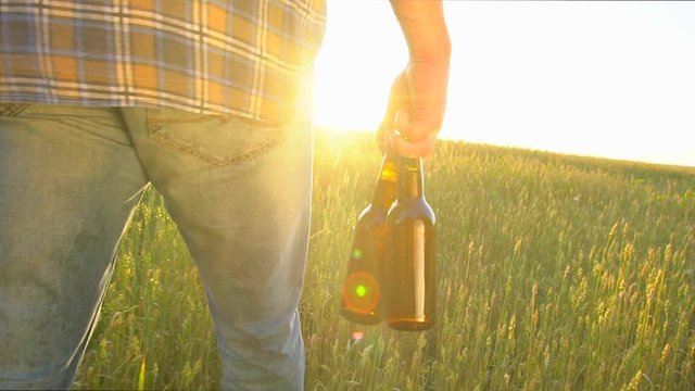 a man walks on a wheat field with two bottles of beer at sunset. Sun lens flare. slow-motion, close-up