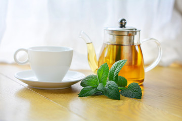 White cup and glass teapot of herbal tea with fresh mint twig on wooden background