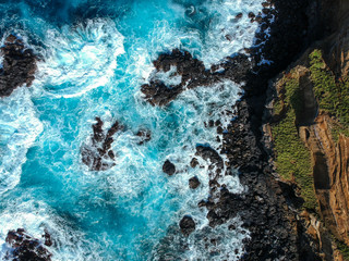 Aerial top view of sea waves hitting black volcanic rocks on the coastline with turquoise sea water. Amazing rock cliff seascape in the Portuguese coastline. Azores islands. Drone shot.