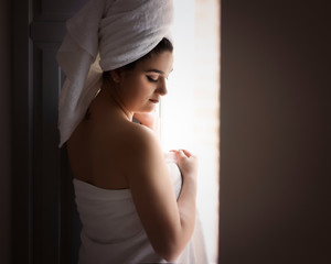 Fototapeta premium Portrait of a beauty girl with a towel in her head and a towel around her body looking through the window