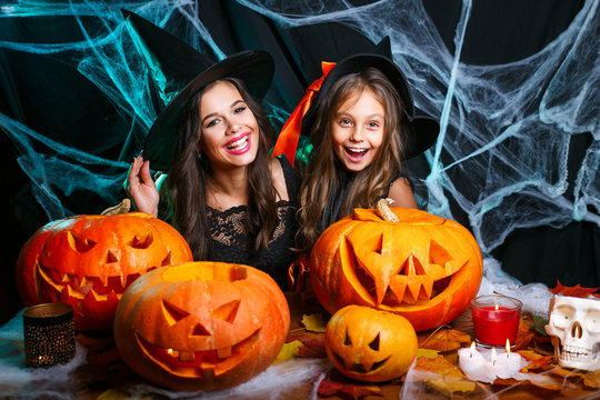 Happy halloween. Cute little girl and her beautiful mom in witch hats are looking at camera and smiling while sitting on wooden background decorated for Halloween