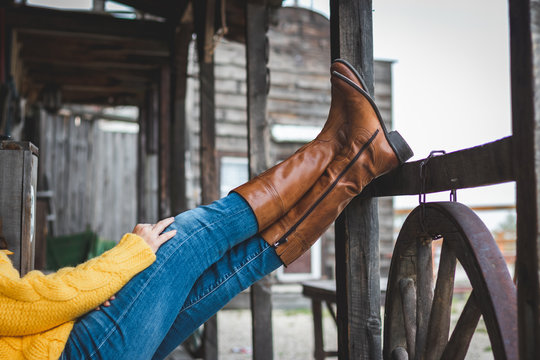 Woman is relaxing on the ranch with her legs on a wooden railing. Leather boot and jeans