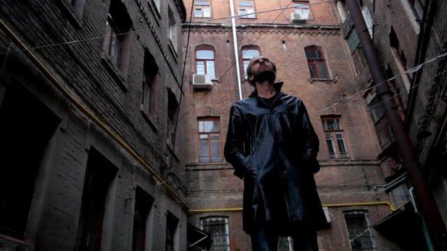 Bearded strange man with black coat stands near old buildings of city