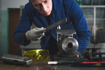 Man worker is working with iron detail on vise grip on blacksmith table and using a file.