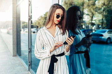 Happy attractive young woman in sunglasses looking at smartphone screen while walking in the city. Dressed in stylish clothes.