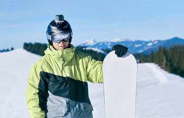 Men snowboarder stands with snowboard on mountain backdrop.