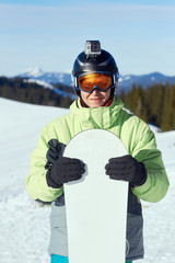 Fototapeta na wymiar .Snowboarder with action camera on a helmet. Portrait of man at the ski resort on the background of mountains and blue sky, hold snowboard. Wearing ski glasses in the mountains