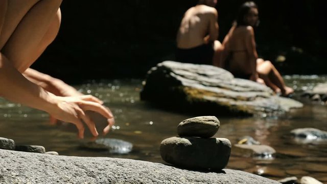 Female puts a cairn close-up standing on a cobblestone in a mountain river against the background of tourists, the tower falls