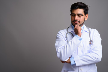 Young handsome bearded Persian man doctor against gray backgroun