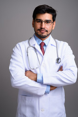 Young handsome bearded Persian man doctor against gray backgroun
