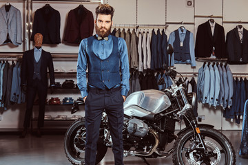 Fototapeta na wymiar Elegantly dressed man posing with hands in pockets near retro sports motorbike at the men's clothing store.