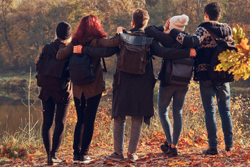 Back view of young friends hugging together and looking at the lake in beautiful autumn forest.