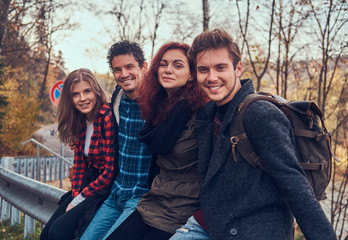 Group of young friends with backpacks sitting on guardrail near road with a beautiful forest and river in the background.