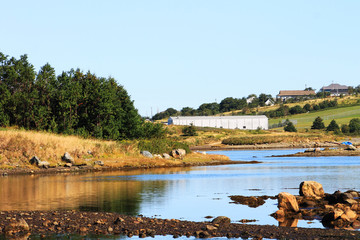 Looking across a shallow harbor at the coastline beyond. An industrial building and some houses are on the shore.