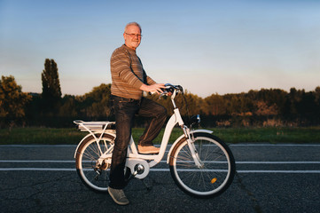 Obraz premium Old man cyclist on the e-bike or electric white bicycle on the nature background. Active pension. Travel. Sport.