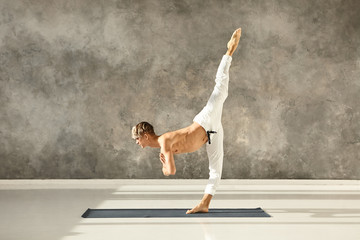 Athletic fit young male with muscular torso wearing no shirt practicing advanced yoga asana, standing with one leg on floor, training balance, concentration and coordination, bending forward - obrazy, fototapety, plakaty