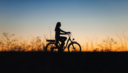 Silhouette of a girl on the e-bike or electric bicycle on the sunset background. Country style. Transportation in the village. Copy space. Female. Travel.