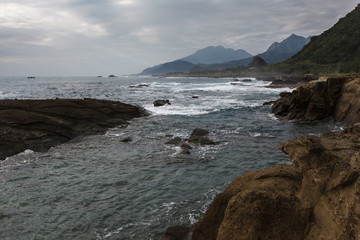 Fototapeta na wymiar Taiwan East Coast Rocky Coastline Background Image - Overcast Skies, Exotic Rock Formations, Grass and Waves in the Ocean. Ocean Coastline, Asia Landscape Photography, Rays of sunshine in background