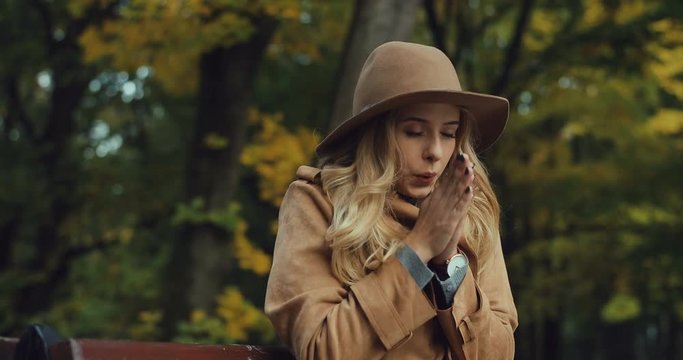 Caucasian good looking young woman feeling cold and trying to warm her hands while she sitting on the bench in the park at fall.