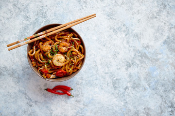 Traditional asian udon stir-fry noodles with shrimp in bowl and chopsticks. Fresh chilli pepers on side. Placed on bright stone background with copy space.