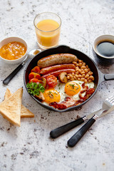 Full English breakfast - fried egg, baked beans, bacon, sausages on a bright rusty background, toasts, orange juice and coffee on a side. Dish in iron frying pan.