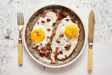 Two fresh fried eggs with crunchy crisp bacon served on rustic plate. Fork and knife on sides. Top view.