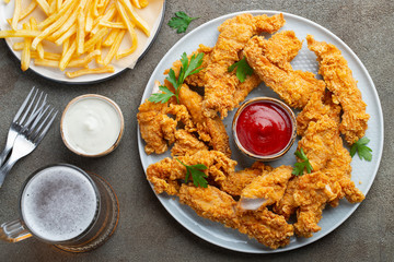 Breaded chicken strips with two kinds of sauces, fried potatoes and beer on a wooden Board. Fast food on dark brown background. Top view