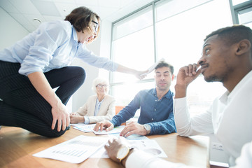 Furious young female employee in shirt crouching on conference table and hitting boss with rolled paper, she expressing her negative emotions