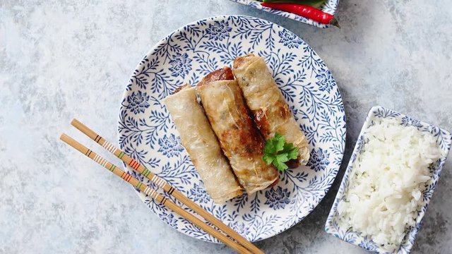 Orginal asian deep fried spring rolls placed on beautiful oriental style plate with chopstick, boiled rice, cucomber nad hot chilli peppers od side. Top view on stone background.
