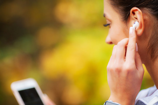 Photo of Joyful Fitness Woman 30s in Sportswear Touching Bluetooth Earpod and Holding Mobile Phone, While Resting in Green Park