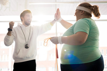 Waist up portrait of smiling fitness coach high fiving with fat young woman during workout in...