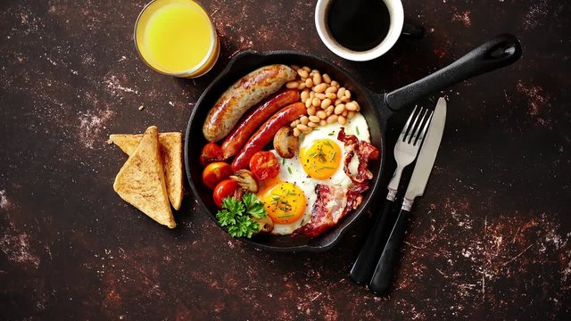 English breakfast in iron cooking pan with fried eggs, sausages, bacon, beaked beans, toasts, orange juice and black coffee on dark rusty background.