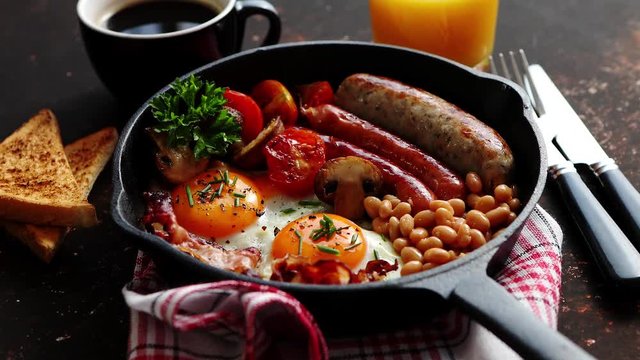 Full English breakfast - fried egg, baked beans, bacon, sausages on a dark rusty background, toasts, orange juice and coffee on a side. Dish in iron frying pan.