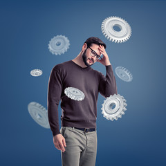 A man in glasses stands deep in thought while being surrounded by several large metal spur gears.