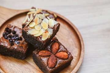Homemade square brownie cake with various topping like almond, nut and chocolate are placing on wooden round plate.