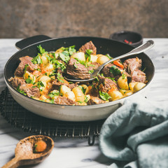 Meat dinner. Braised beef meat with potato and carrot with fresh parsley in cooking pan over white marble table background, selective focus, copy space, square crop. Comfort winter food