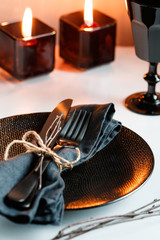 Festive table setting in a black style among black candles on a white table. Plate with fork and knife on a linen napkin. Thanksgiving or Halloween dinner.