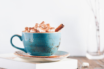 Hot chocolate with marshmallows and cinnamon stick in a blue ceramic cup on a table with a book. The concept of winter or fall time. Minimal scandinavian design.