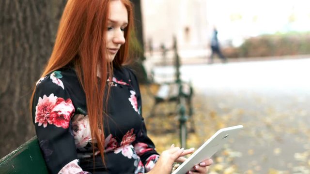 Young pretty woman with red hair working with tablet on the bench park in the city, 4K
