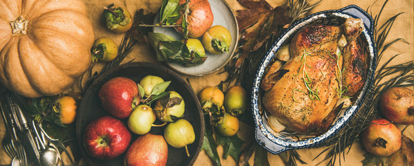 Thanksgiving dinner table. Flat-lay of roasted chicken or turkey, fruit, pumpkin, cutlery, leaves over yellow table runner on grey concrete background, top view, wide composition