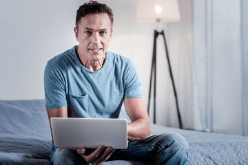 Im busy. Serious occupied confident man sitting in the bedroom looking straight and using the laptop.