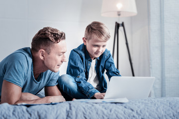 You can do this. Concentrated interested little boy sitting on the bed near his father and using the laptop and looking at the screen.