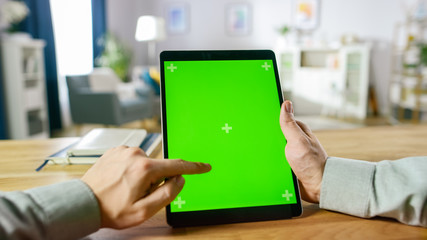 Close-up of Man Using Hand Gestures on Green Mock-up Screen Digital Tablet Computer in Portrait Mode while Sitting at His Desk. In the Background Cozy Living Room.