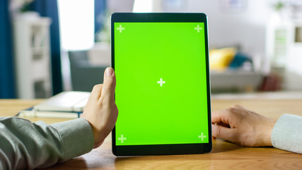 Close-up of Man Using Hand Gestures on Green Mock-up Screen Digital Tablet Computer in Portrait Mode while Sitting at His Desk. In the Background Cozy Living Room.