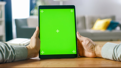 Close-up of Man Using Green Mock-up Screen Digital Tablet Computer in Portrait Mode while Sitting at His Desk. In the Background Cozy Living Room.