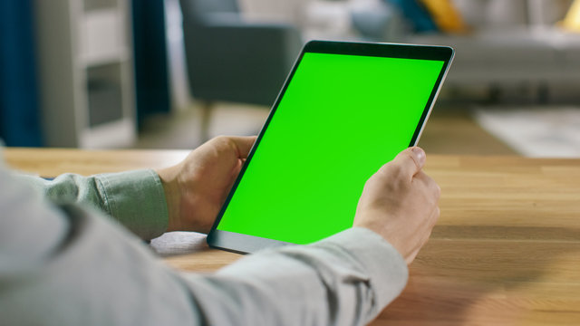 First Person Shot of Man Using Hand Gestures on Green Mock-up Screen Digital Tablet Computer in Portrait Mode while Sitting at His Desk. In the Background Cozy Living Room.