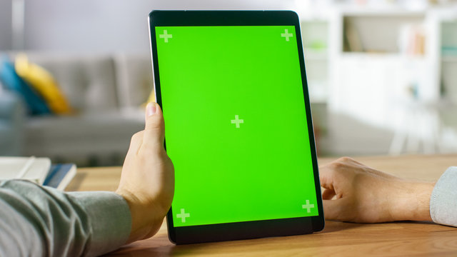 Close-up of Man Using Hand Gestures on Green Mock-up Screen Digital Tablet Computer in Portrait Mode while Sitting at His Desk. In the Background Cozy Living Room.